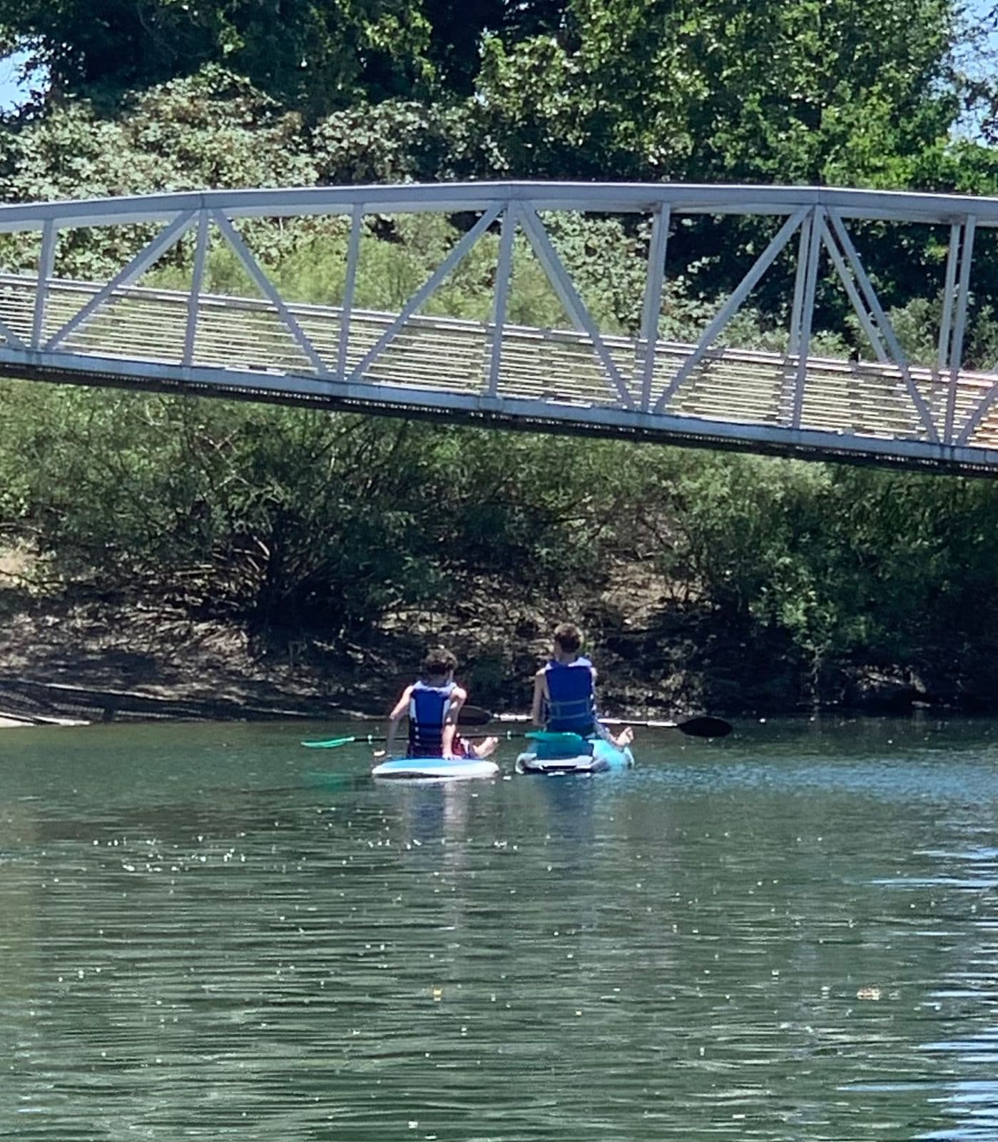 Two paddlers in life vests sit on hybrid boards, paddling away from the camera on calm water and passing under a small white footbridge lined with trees