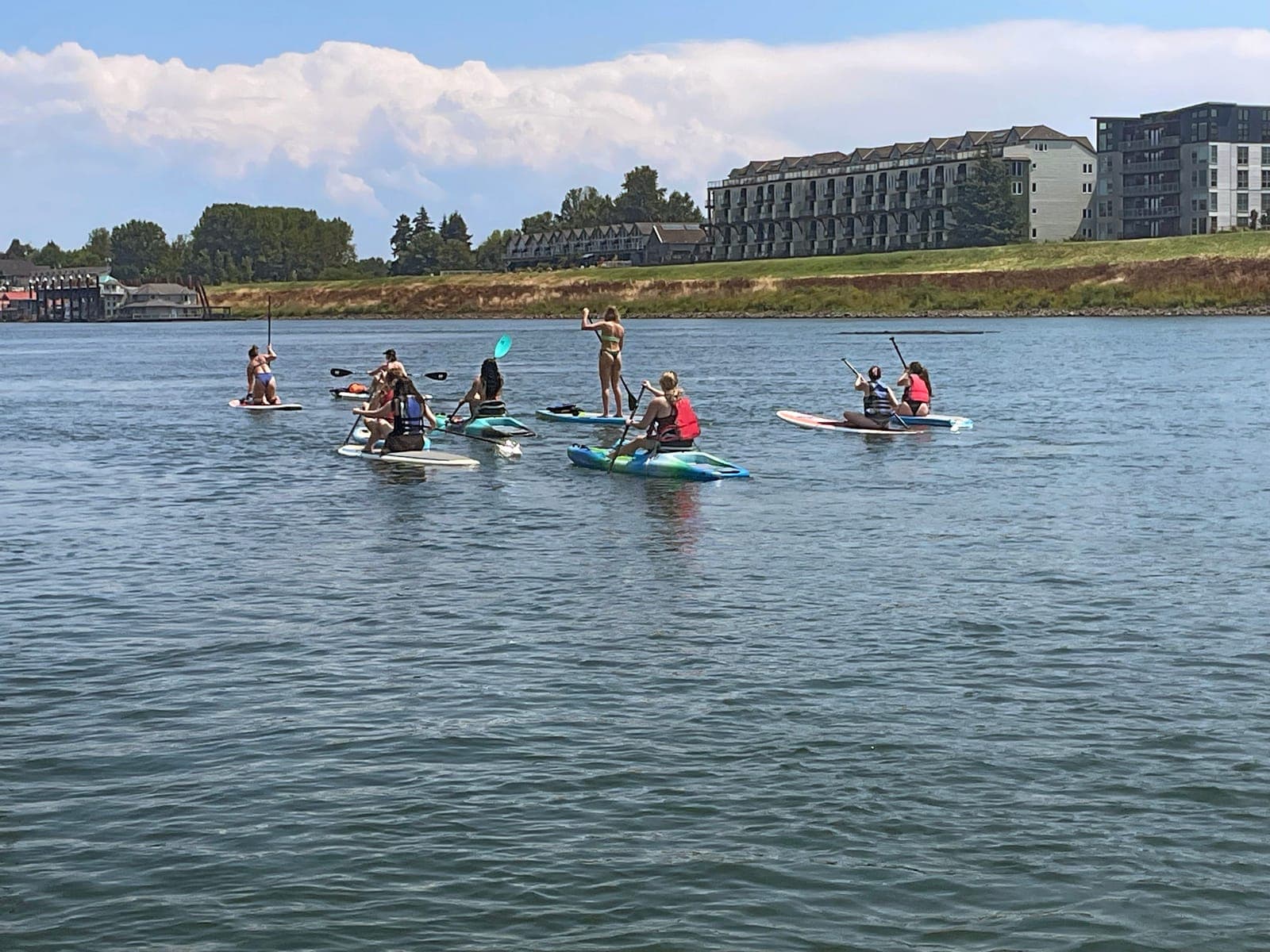 A group of paddlers on hybrid boards, some standing and some sitting, wearing life vests on the water