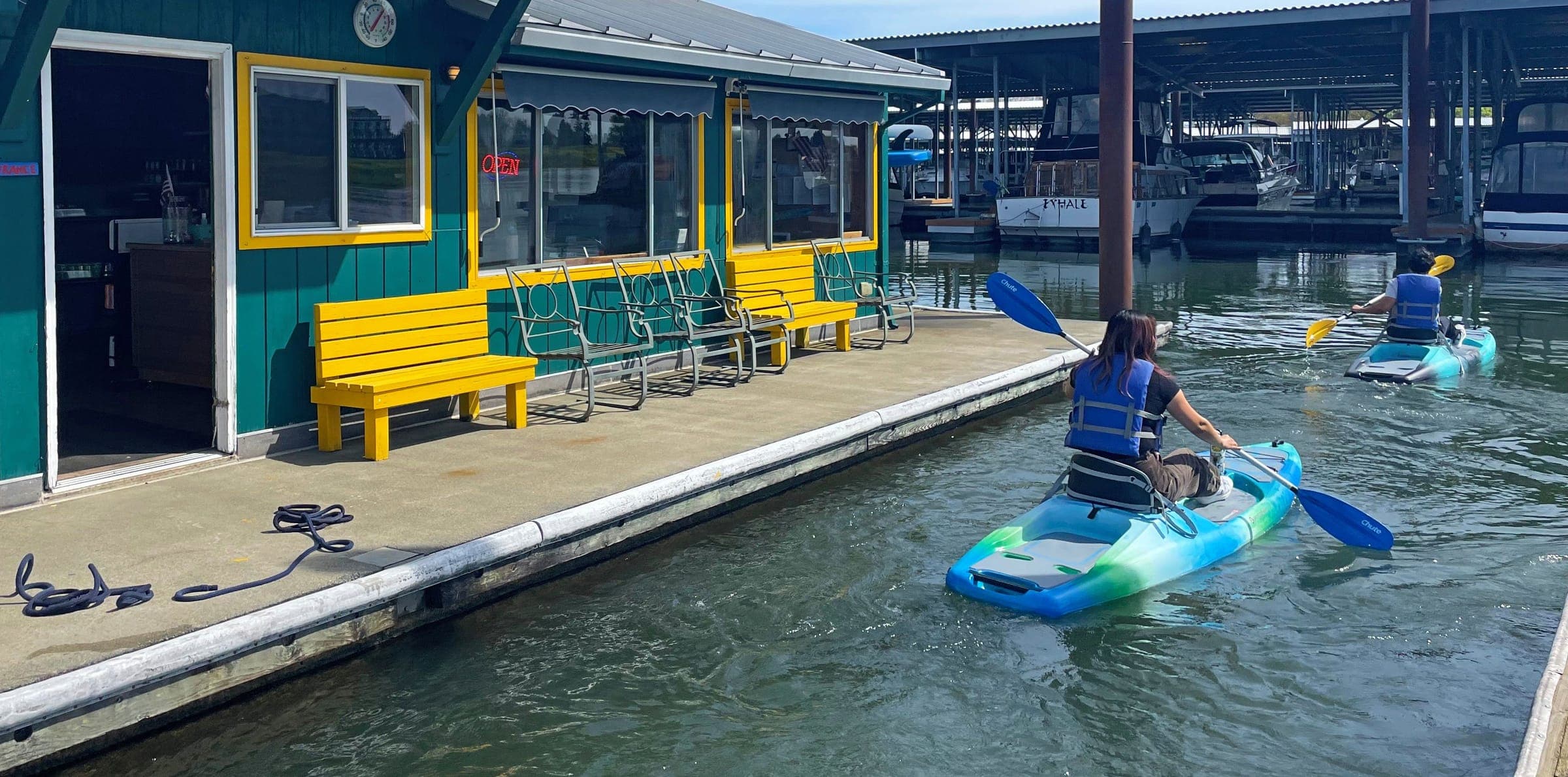 Two kayakers in life vests paddling away from the green-and-yellow dock store into the marina