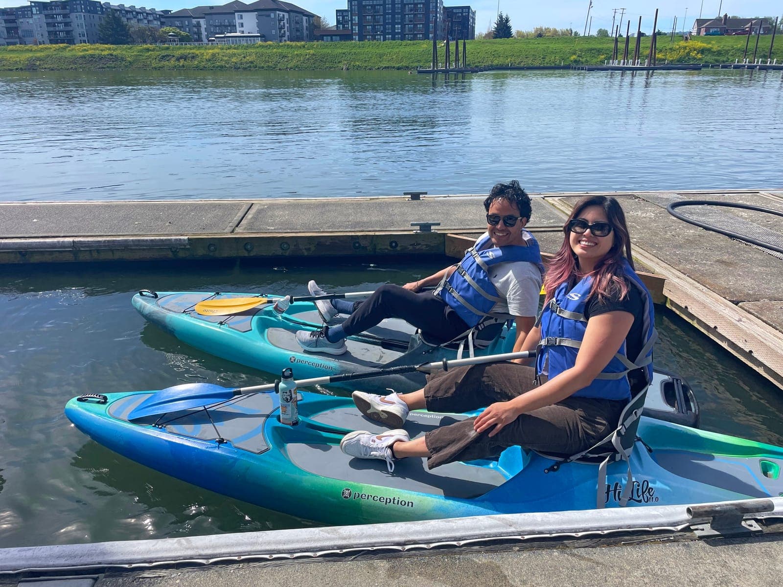Two smiling paddlers in life vests seated on hybrid kayaks at the dock, ready to go