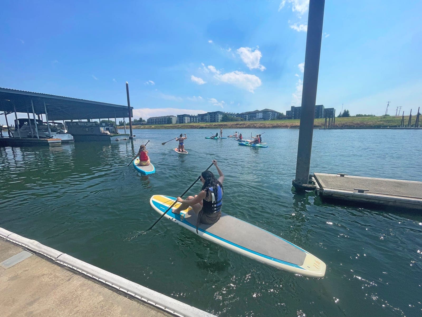 A paddleboarder kneeling on a board pushes off from the dock into the marina, with other paddlers ahead