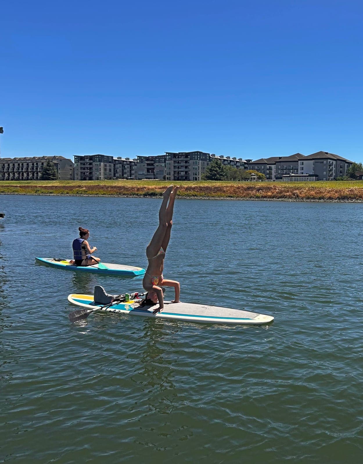 A paddler does a headstand on a paddleboard on calm water, with another paddler nearby